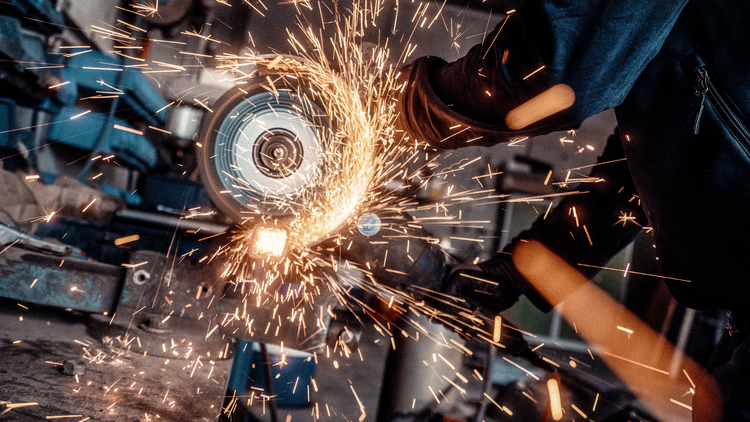 Eine Person trägt Handschuhe und benutzt eine Maschine, die Funken sprüht. Die Umgebung ist schattig, und es sind Maschinen und Werkzeuge im Hintergrund sichtbar. Die Szene vermittelt eine Atmosphäre industrieller Arbeit und Handwerk.