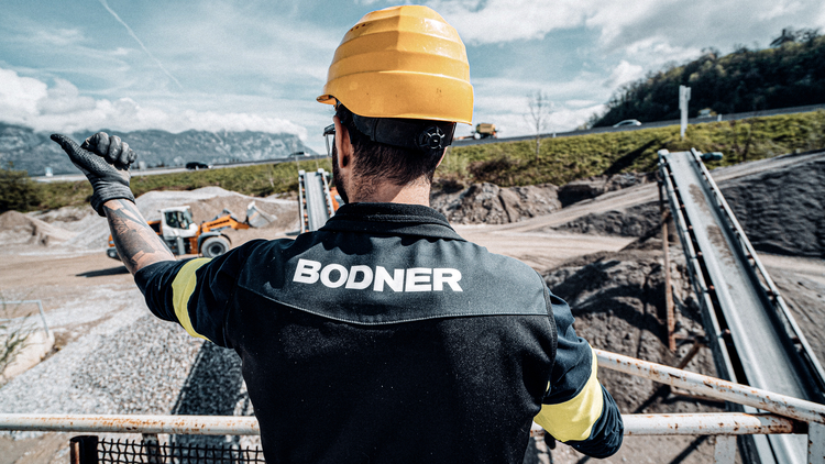 Ein Arbeiter mit gelbem Helm und schwarzer Arbeitskleidung steht auf einer Baustelle. Er zeigt mit einer Hand in Richtung einer Maschinenanlage im Hintergrund, während Schotter und ein Bagger sichtbar sind. Die Umgebung ist von Bergen und leicht bewölktem Himmel umgeben.