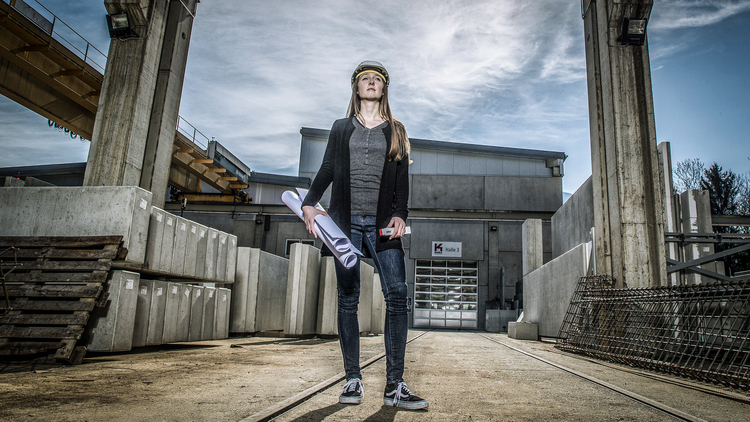 Eine selbstbewusste Frau steht in einer Baustellenumgebung. Sie trägt einen Helm und hält Baupläne in der Hand. Die Kulisse besteht aus Betonblöcken und einem Gebäude im Hintergrund. Der Himmel ist bewölkt, was auf eine arbeitsreiche Atmosphäre hinweist.