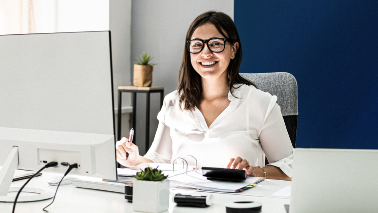 Eine junge Frau sitzt an einem Schreibtisch in einem modernen Büro. Sie trägt eine weiße Bluse und Brille, lächelt freundlich und hält einen Stift in der Hand. Vor ihr liegen Unterlagen, ein Taschenrechner und eine kleine Pflanze auf dem Tisch.