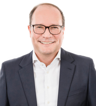 Thomas Bodner - Porträt eines lächelnden Mannes mit Brille, der eine dunkle Jacke und ein weißes Hemd trägt. Der Mann wirkt freundlich und professionell.