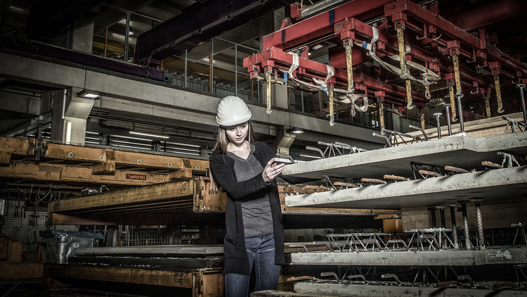 Eine junge Frau mit einem Schutzhelm stehend in einer Fabrikhalle. Sie schaut auf ihr Smartphone und ist umgeben von Betonplatten mit Stahlverstärkung. Im Hintergrund sind große Maschinen und einen Kran zu sehen, der wohl mit den Platten arbeitet.