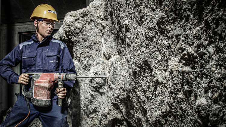 Ein Arbeiter in einem blauen Arbeitsanzug und einem gelben Helm hält einen schweren Bohrer. Er steht vor einer rauen Betonwand, während er die Maschine bereit macht, um in das Material einzudringen. Staub ist sichtbar, was auf die Arbeit hinweist.